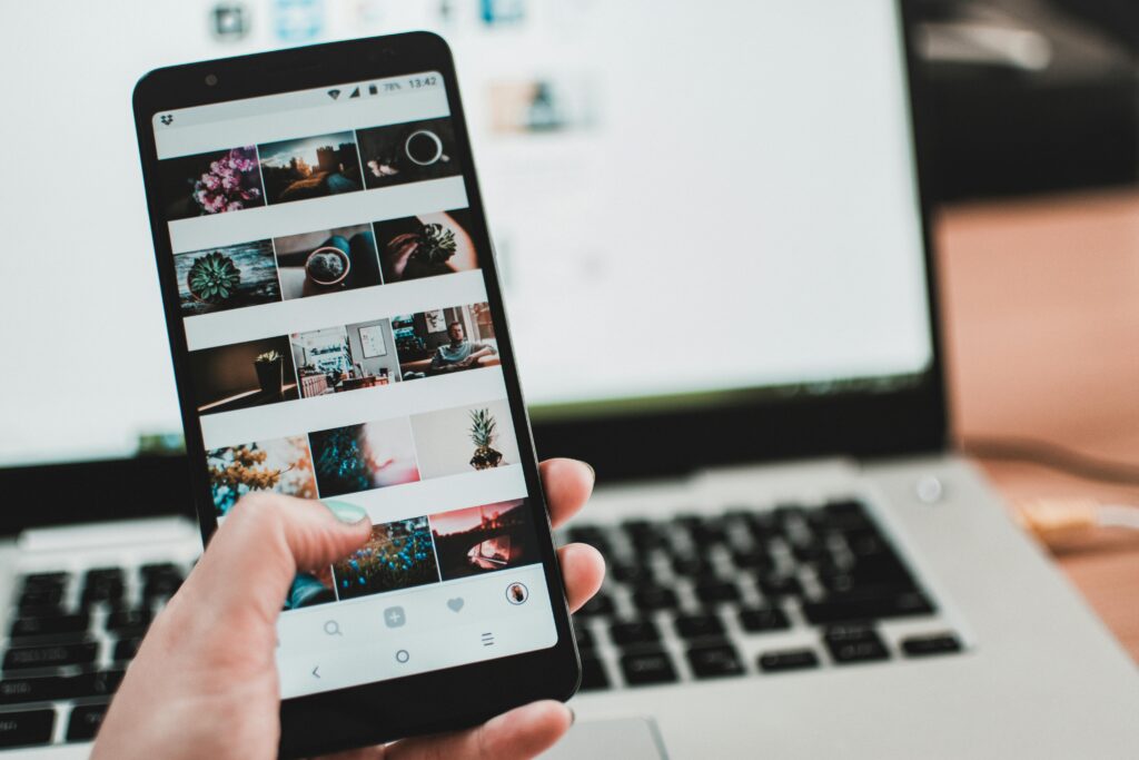 A person uses a smartphone to browse social media in front of a laptop, emphasizing modern technology and lifestyle.